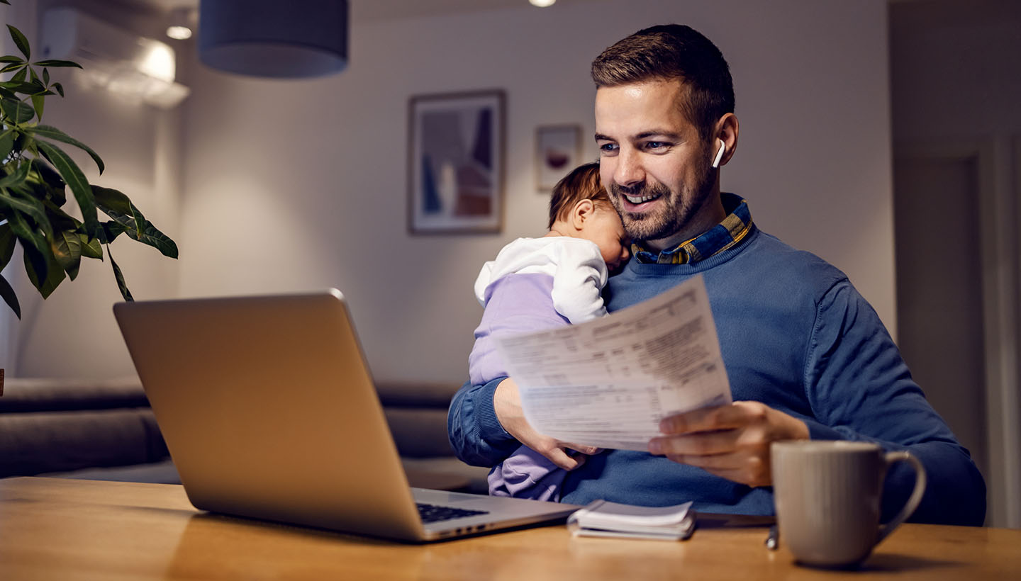 Man looking at computer with a document in one hand and a baby sleeping on his shoulder. Presumably shopping benefits coverage.