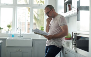 A home inspector standing in a bright kitchen, reading a document thoughtfully while leaning against the counter.