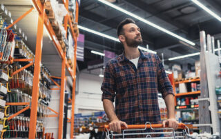 Man in a plaid shirt pushing a shopping cart through a hardware store aisle, surrounded by tools and equipment on shelves