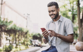 Smiling man sitting outdoors, using a smartphone while wearing a light-colored shirt and jeans in a park-like urban setting
